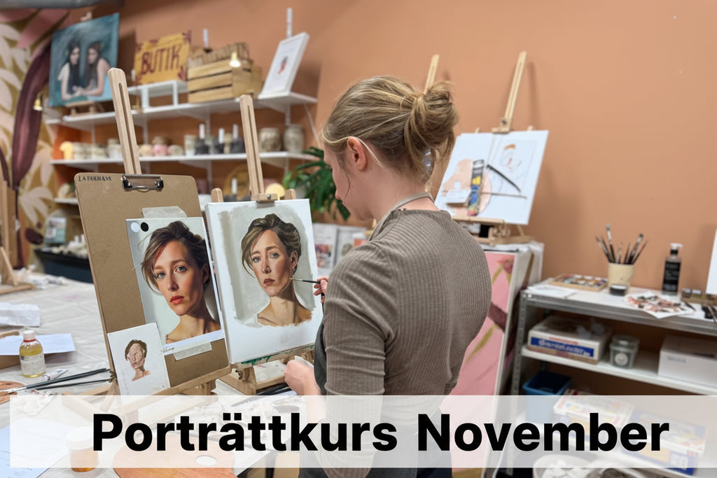 A woman in a gray apron stands at an easel in a portrait painting studio, working on a realistic face painting based on a reference photo. The easel displays the painted portrait alongside the original reference photo, with paint palettes and brushes on a table in the foreground.